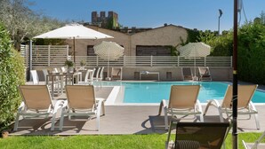 A pool area with lounge chairs and umbrellas, a brick building in the background, and a clear blue sky. - Vialeromadodici Rooms & Apartments (Lazise)