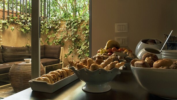 A dining area with a table set for breakfast, including pastries and fruits, and a view of a lush garden through a glass door. - Vialeromadodici Rooms & Apartments (Lazise)