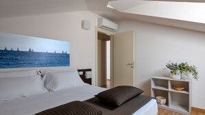 A hotel room with a bed, a painting of a coastal scene, a white air conditioning unit, a wooden floor, and a small plant on a shelf. - Vialeromadodici Rooms & Apartments (Lazise)