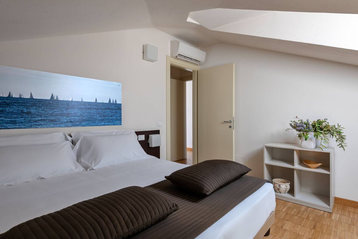 A hotel room with a bed, a painting of a coastal scene, a white air conditioning unit, a wooden floor, and a small plant on a shelf.