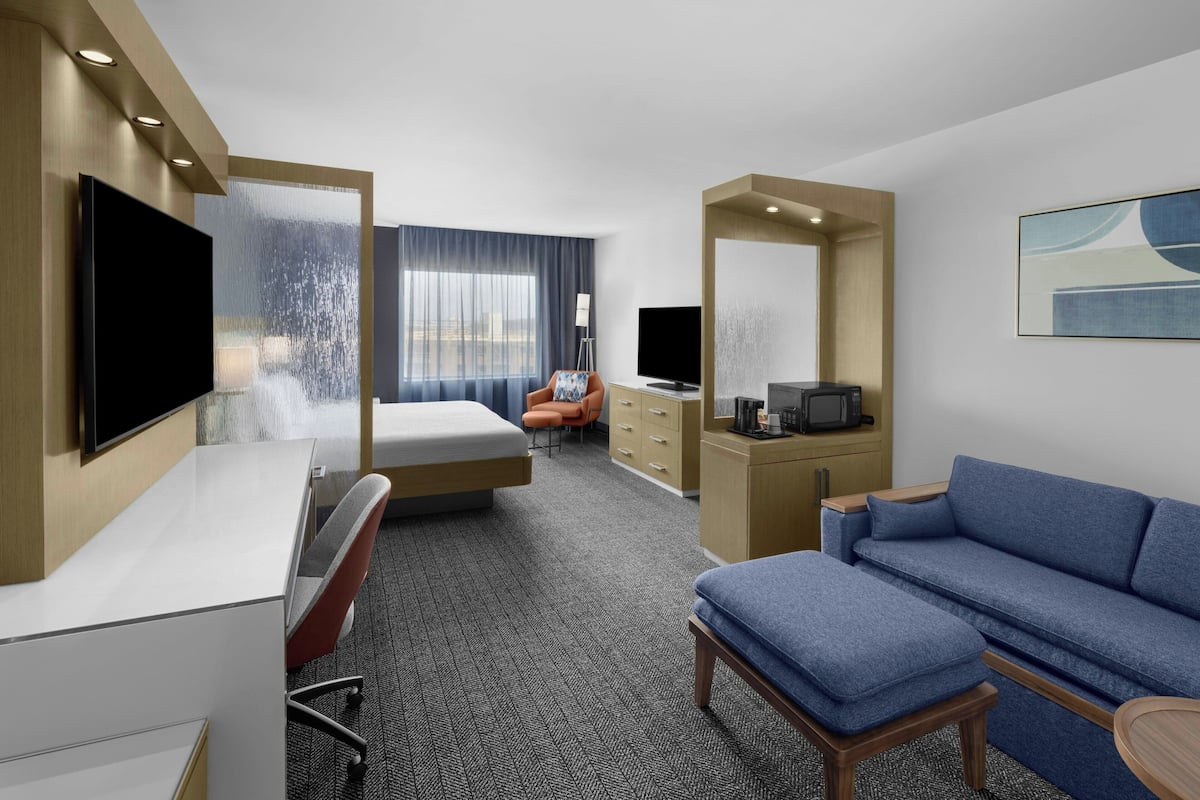 Una habitación de hotel con cama, escritorio, sofá azul, televisor y ventana grande con cortinas.