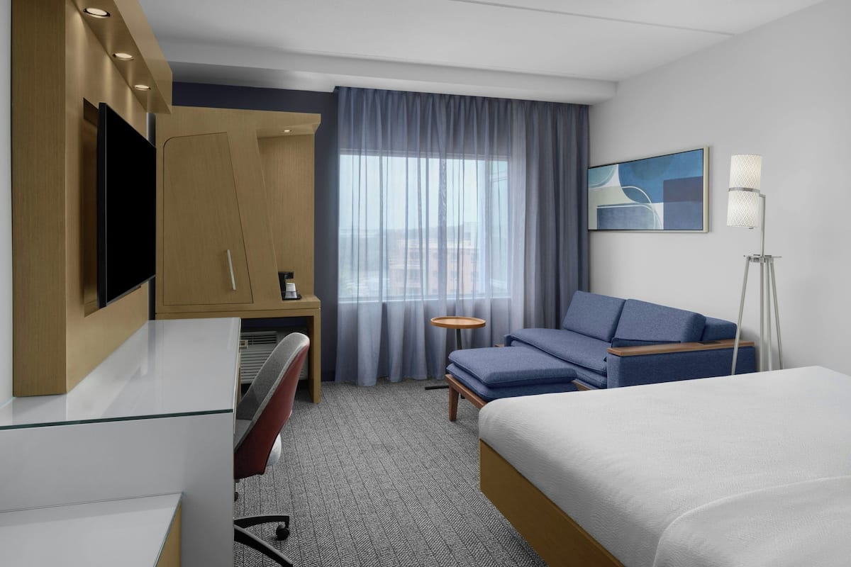 Una habitación de hotel moderna con cama, escritorio, silla, sofá, televisor, lámpara y ventana con cortinas.
