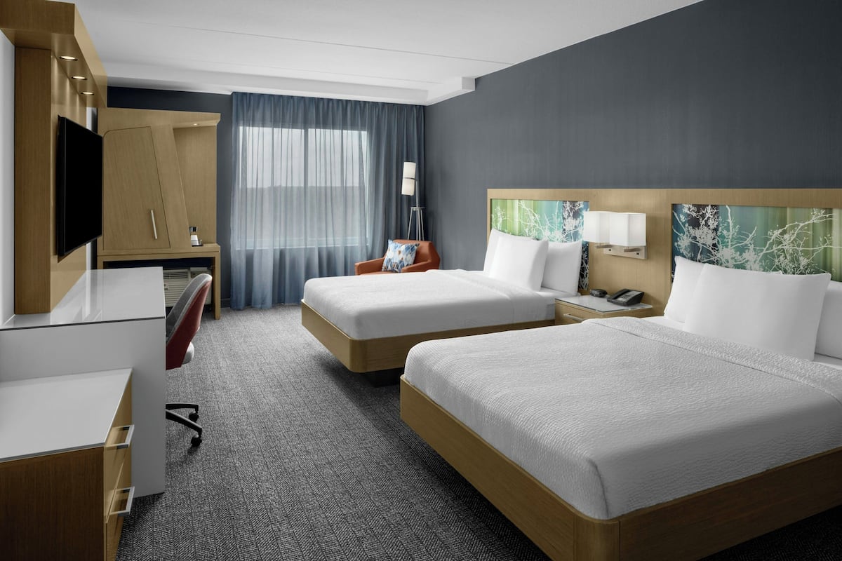Habitación de hotel con dos camas, un escritorio, una silla, un televisor y un ventanal grande con cortinas.