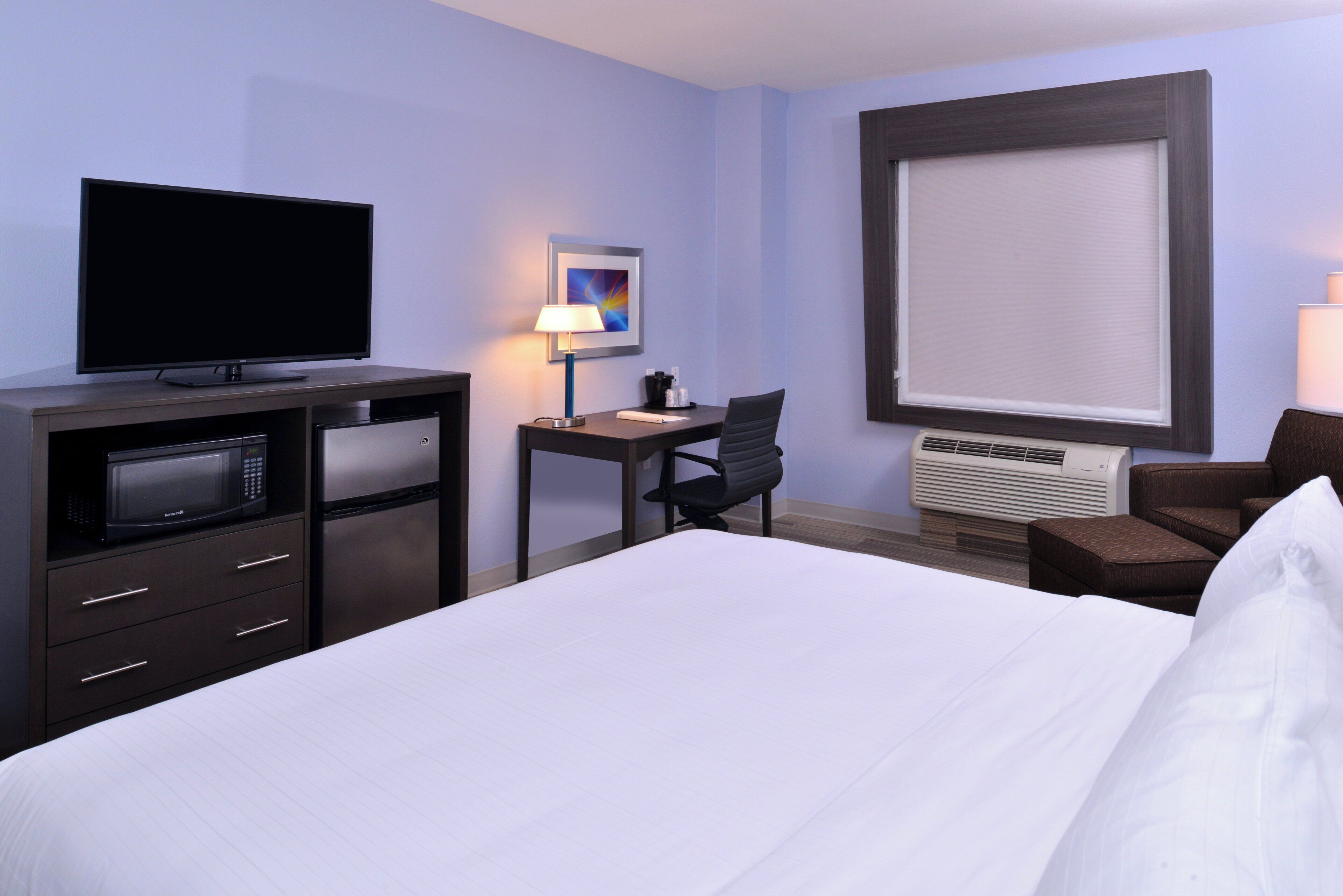 A hotel room with a bed, a desk, a chair, a television, a lamp, and an air conditioning unit.
