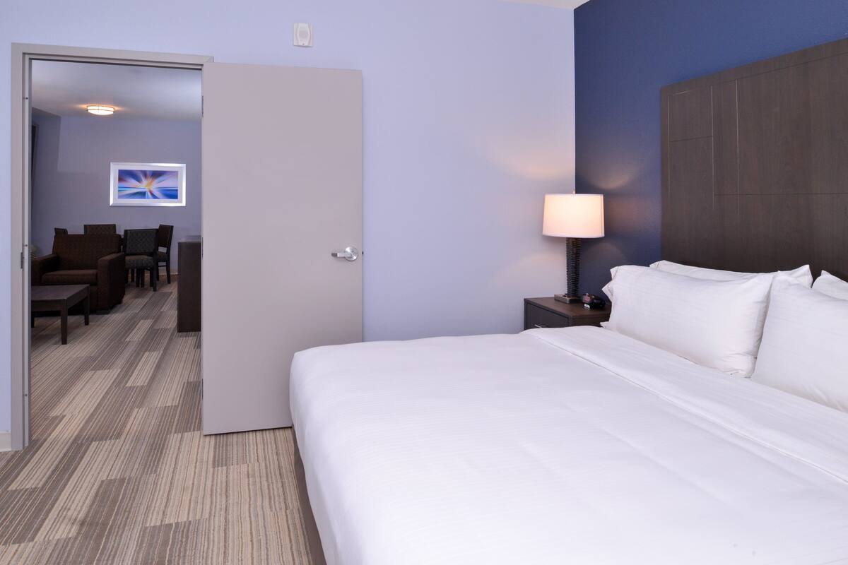A hotel room with a large bed, a bedside lamp, a desk with a chair, and a view of a hallway with a painting.