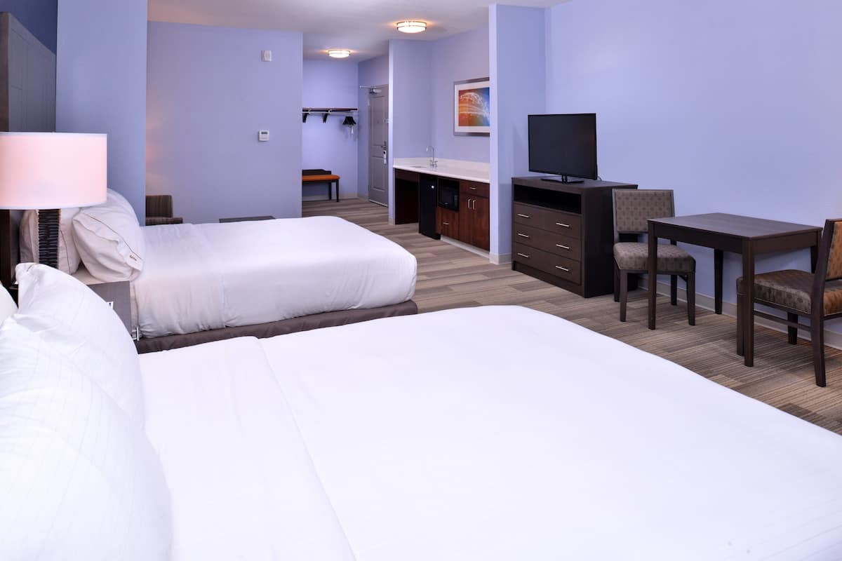 A hotel room with two beds, a desk, a chair, a television, and a small kitchenette.