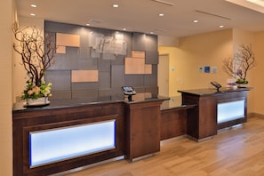 Zona de recepción de un hotel con dos mostradores, diseño moderno con paneles de madera y una superficie de vidrio, y un quiosco de registro digital.