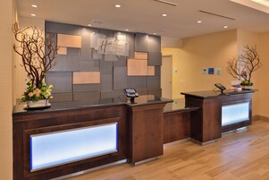 Uma área de recepção de hotel com dois balcões, design moderno com painéis de madeira e tampo de vidro, e um quiosque de check-in digital.