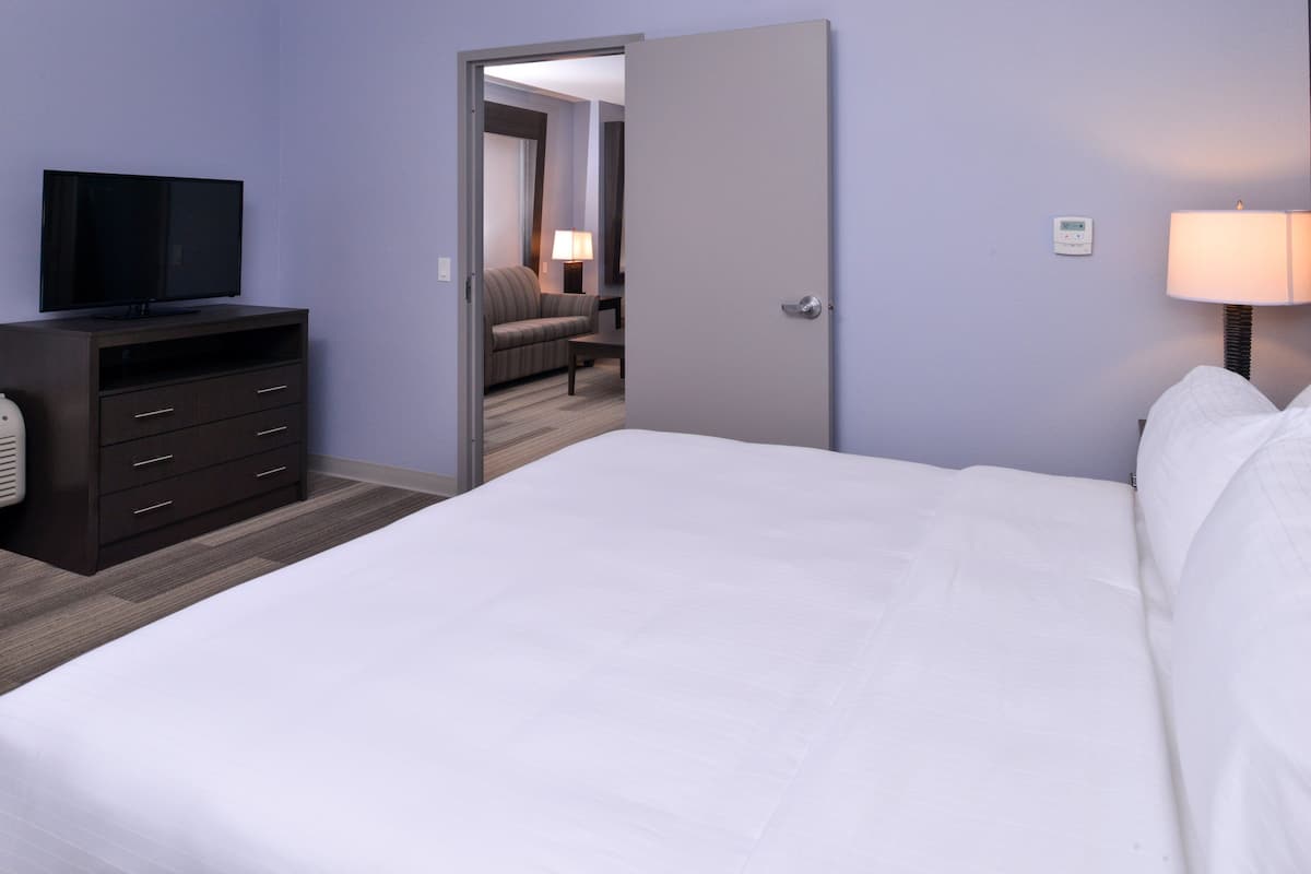 A hotel room with a bed, a TV on a dresser, a lamp, and a door leading to a sitting area.