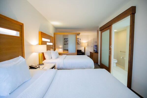 A hotel room with two beds, a desk, a chair, a mirror, a bathroom with a toilet and shower, and wooden-framed sliding doors.