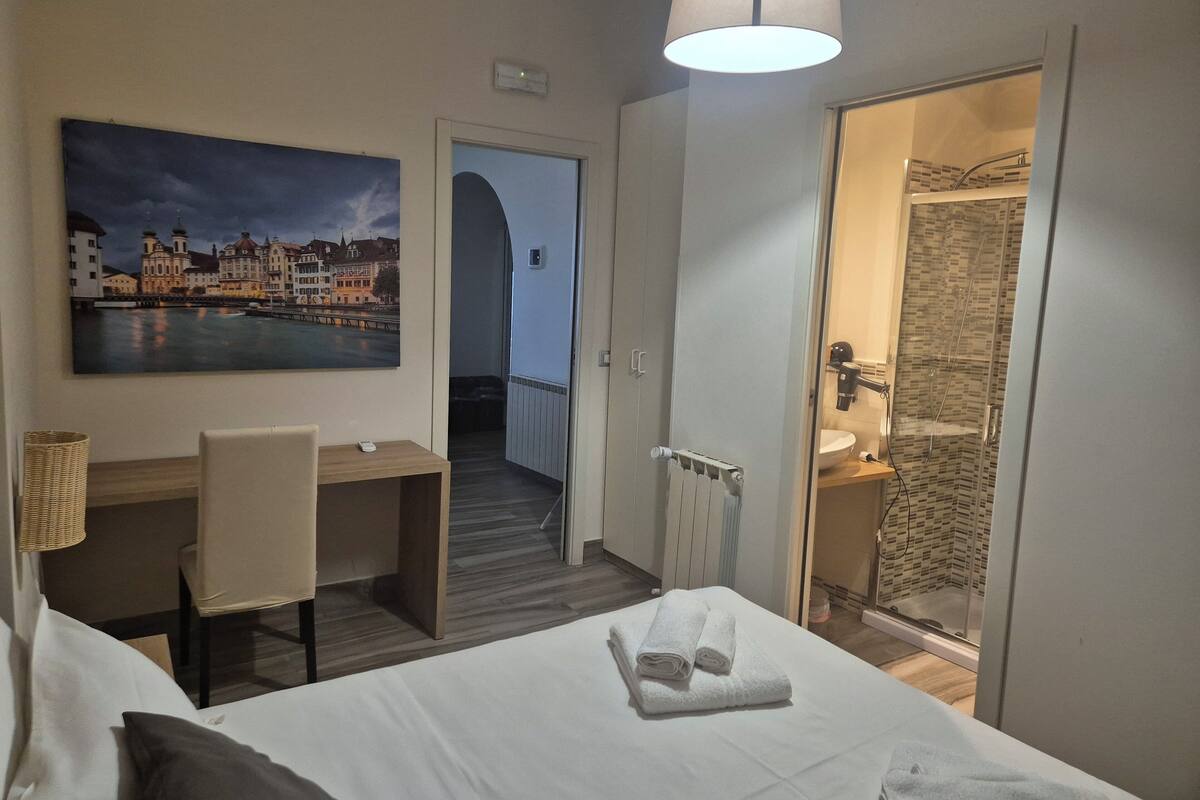 A hotel room with a bed, a desk, a chair, a painting of a cityscape, and a shower area.