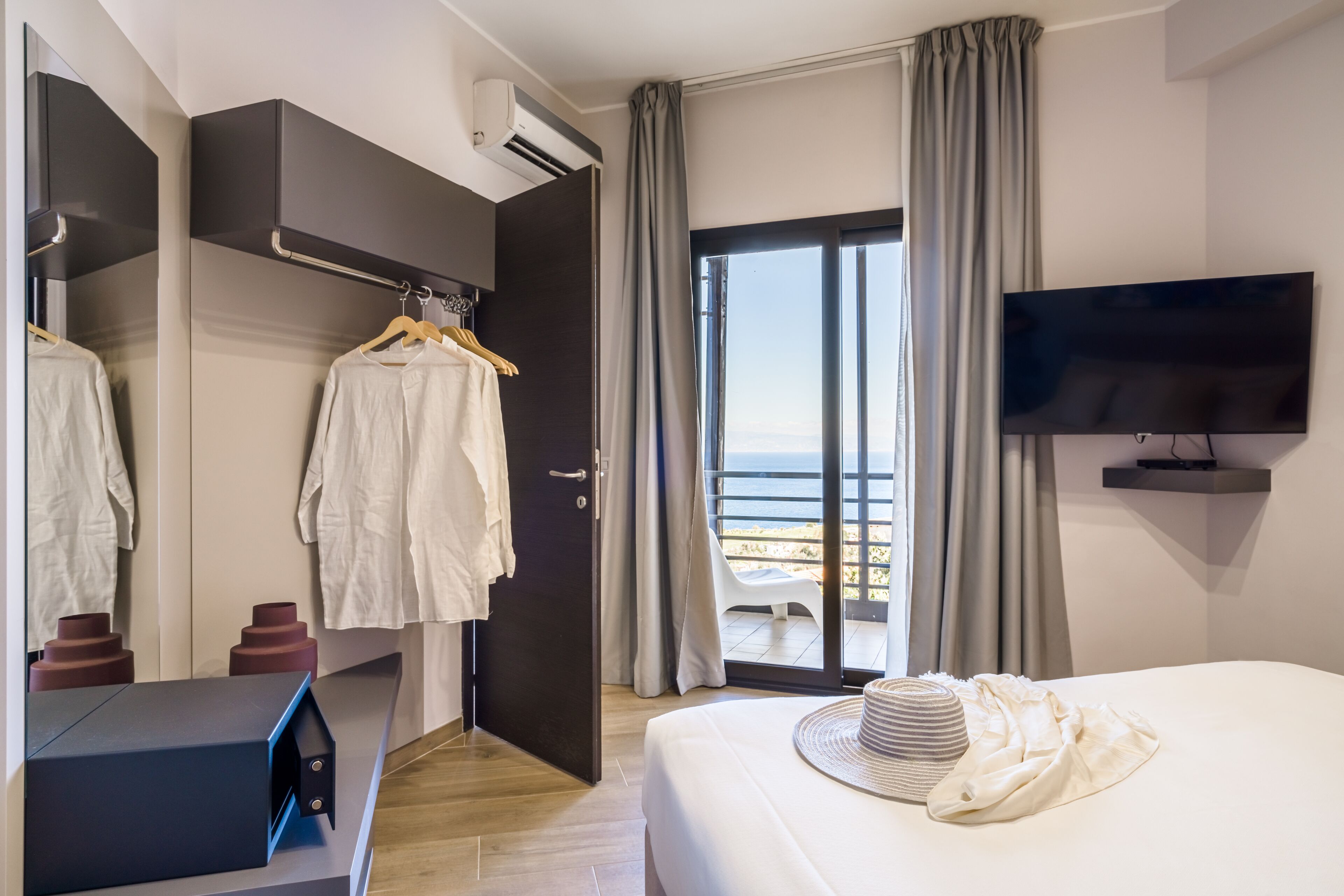 A hotel room with a bed, a TV, a wardrobe, a mirror, and a view of the sea through a sliding glass door.