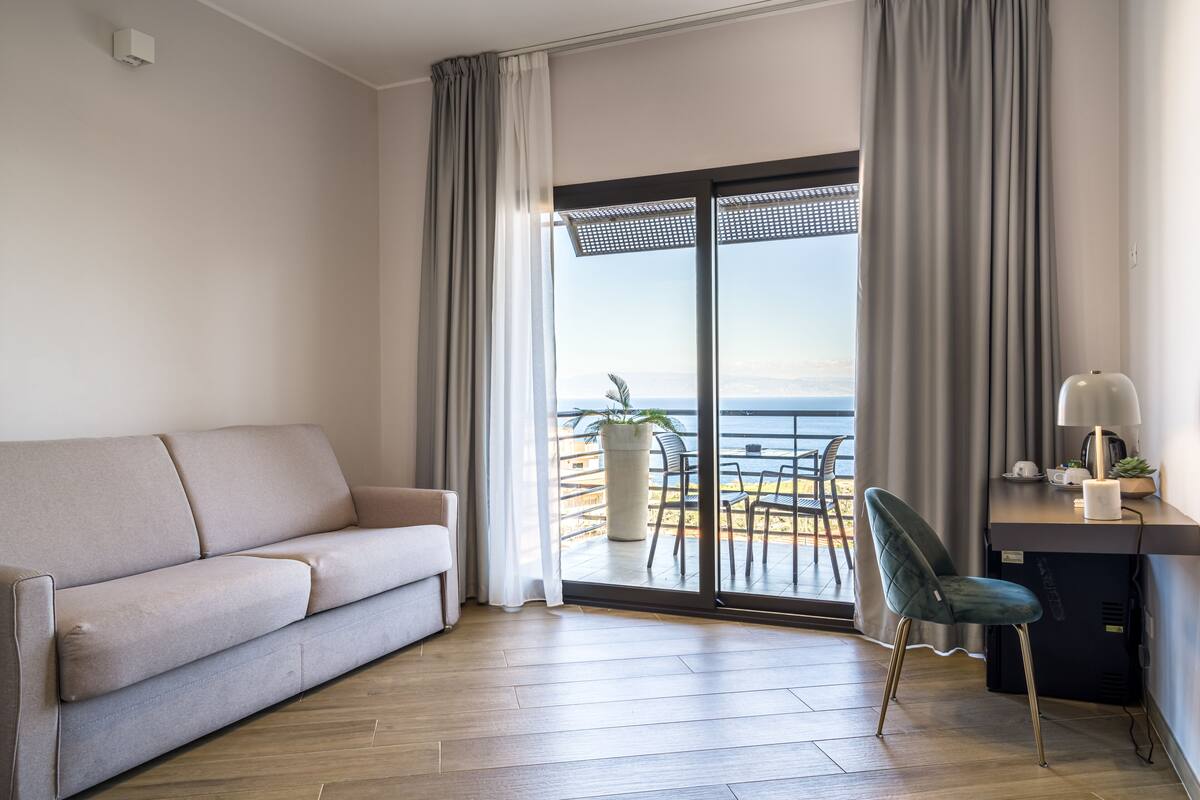 A modern living room with a sofa, a desk with a chair, and a view of the sea through a sliding glass door.