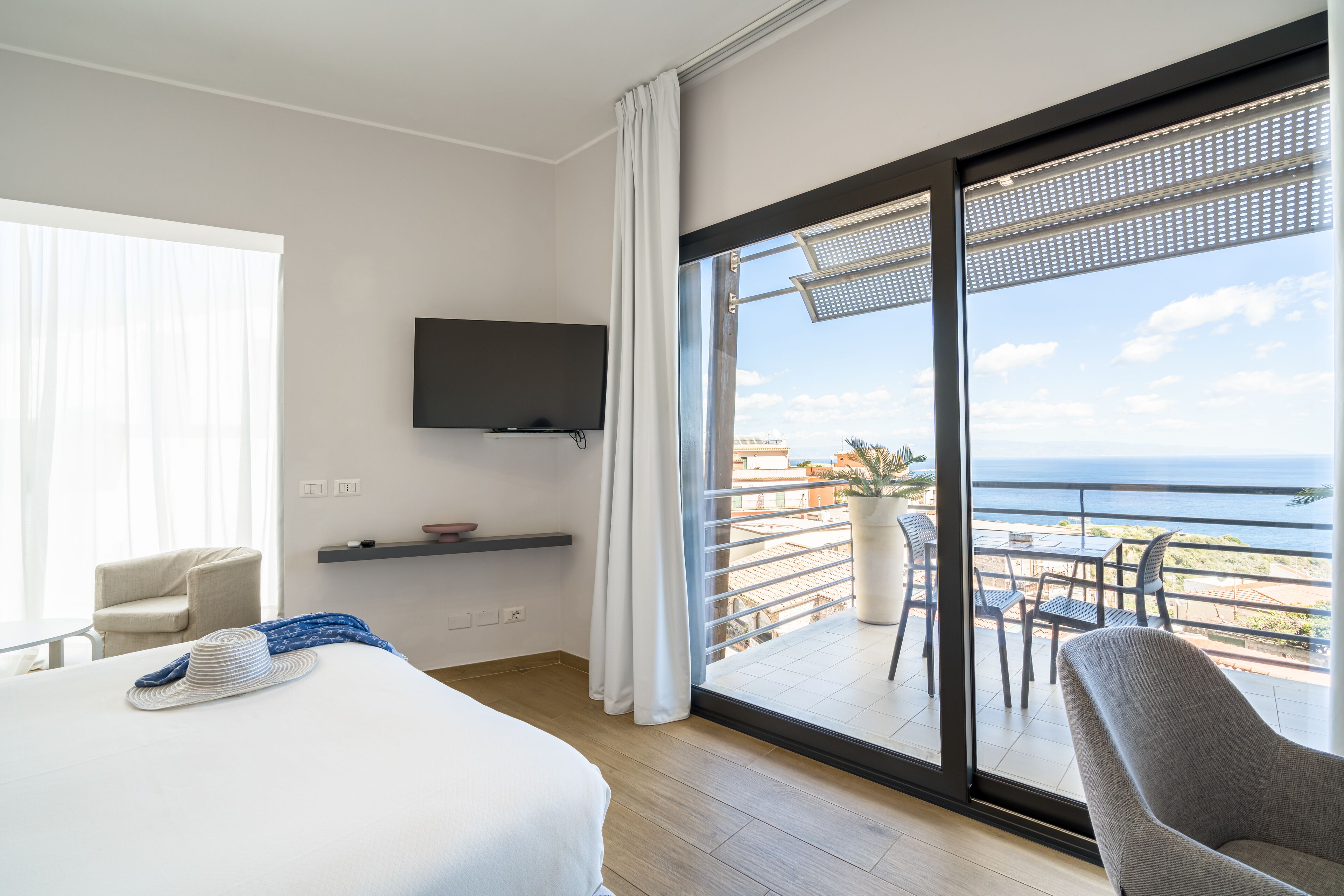 Habitación de hotel con una cama grande, televisor montado en la pared, balcón con vista al mar y una silla.