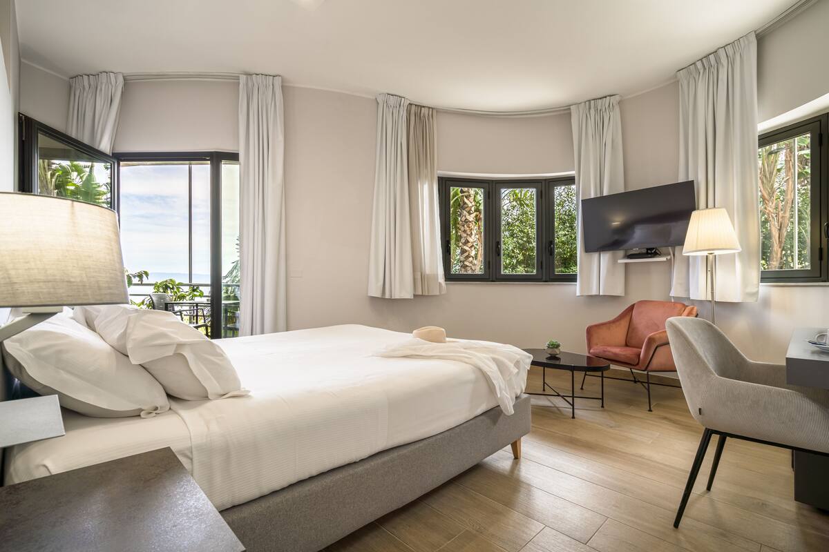 A hotel room with a bed, a desk, a chair, a TV, and large windows with curtains.