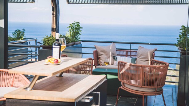 Una terraza con mobiliario de mimbre, una mesa con frutas y una botella, y vista al mar.