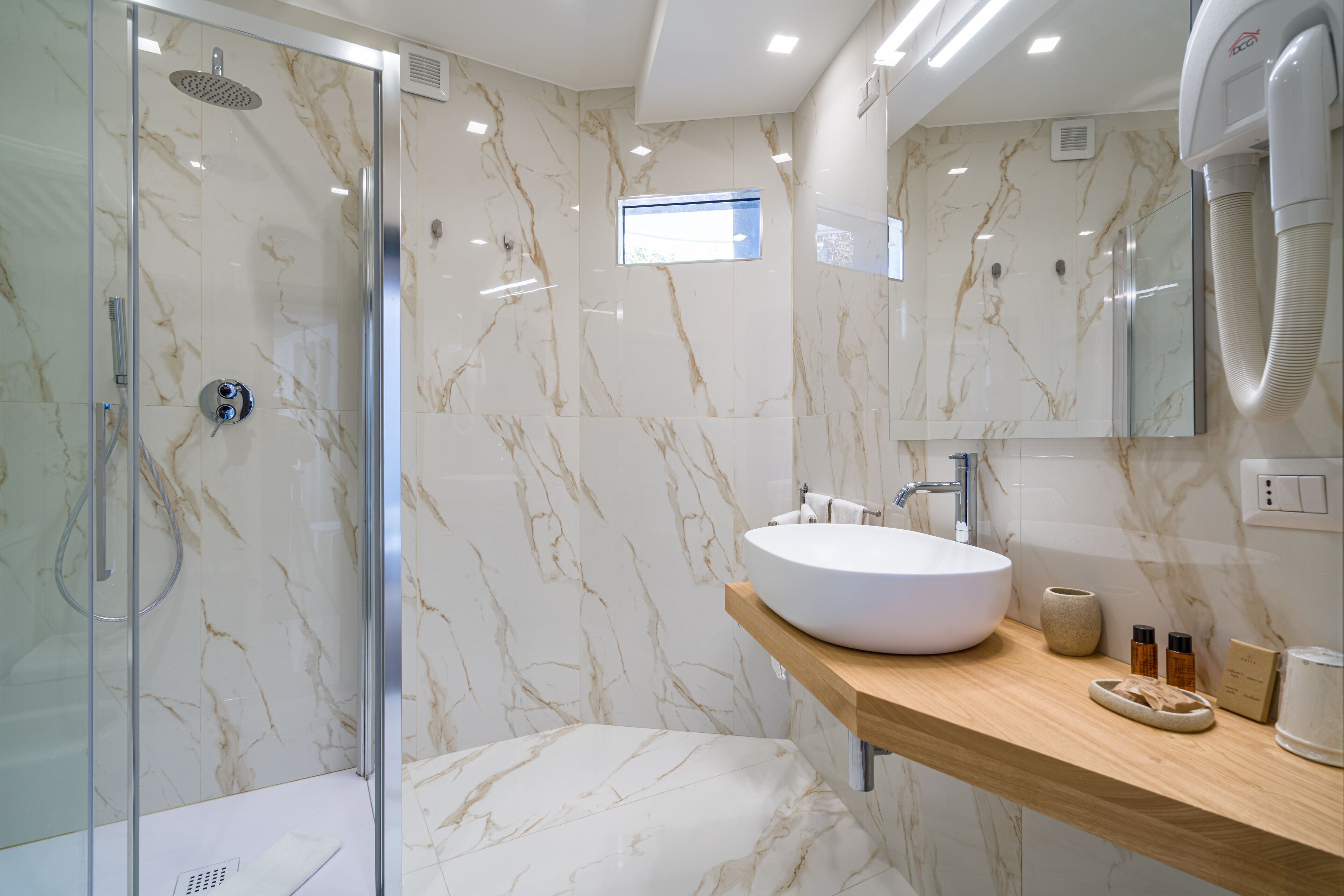 A modern bathroom with a glass-enclosed shower, a marble-tiled wall, a wooden vanity with a white sink, and a handheld showerhead.