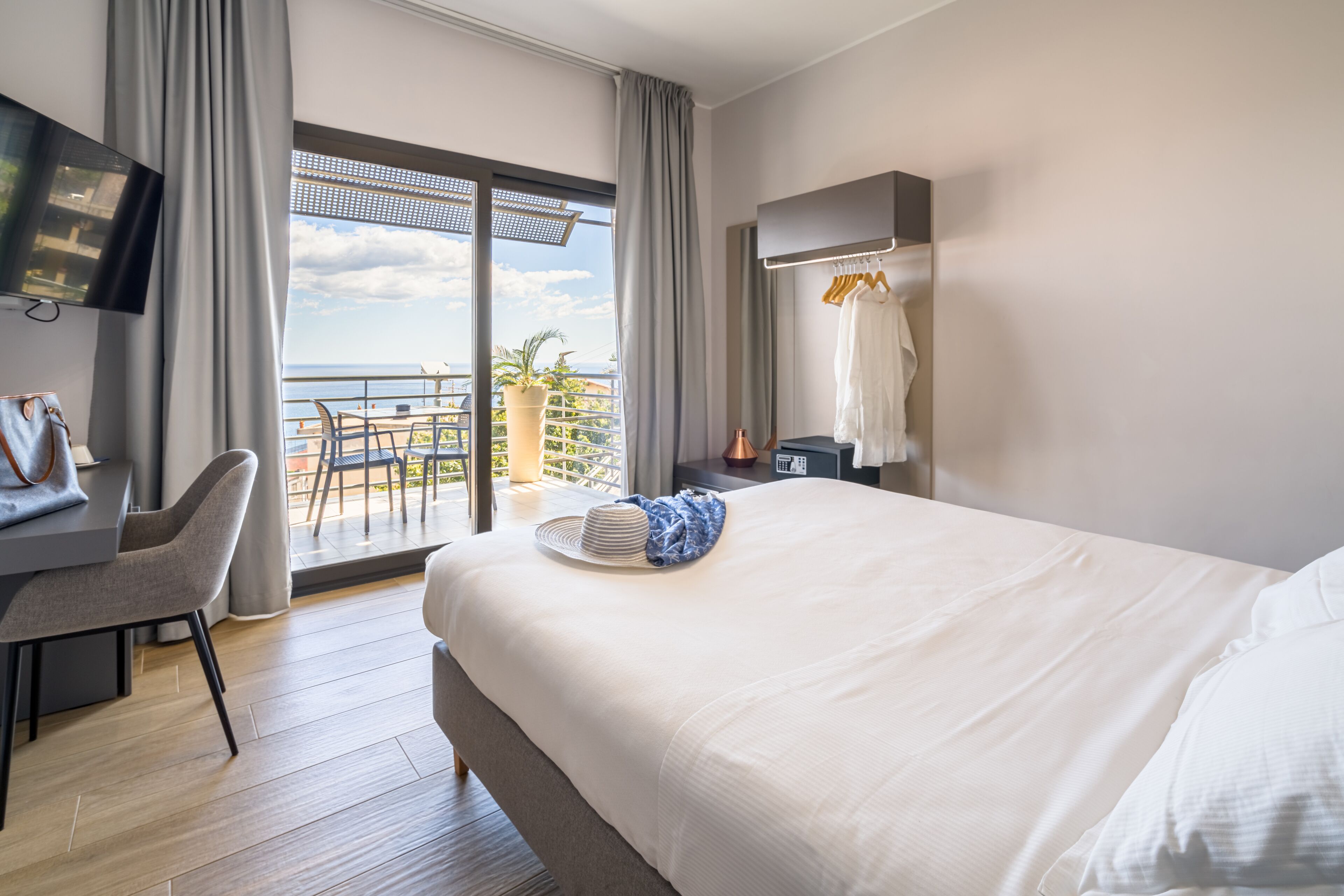 A hotel room with a large bed, a desk with a chair, a TV, and a balcony with a view of the sea.