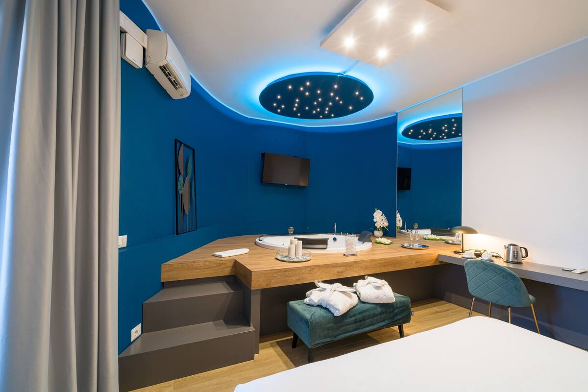 A modern bedroom with a blue accent wall, a wooden vanity, a round mirror, a flat-screen TV, a bedside table with a lamp, and a green upholstered bench.