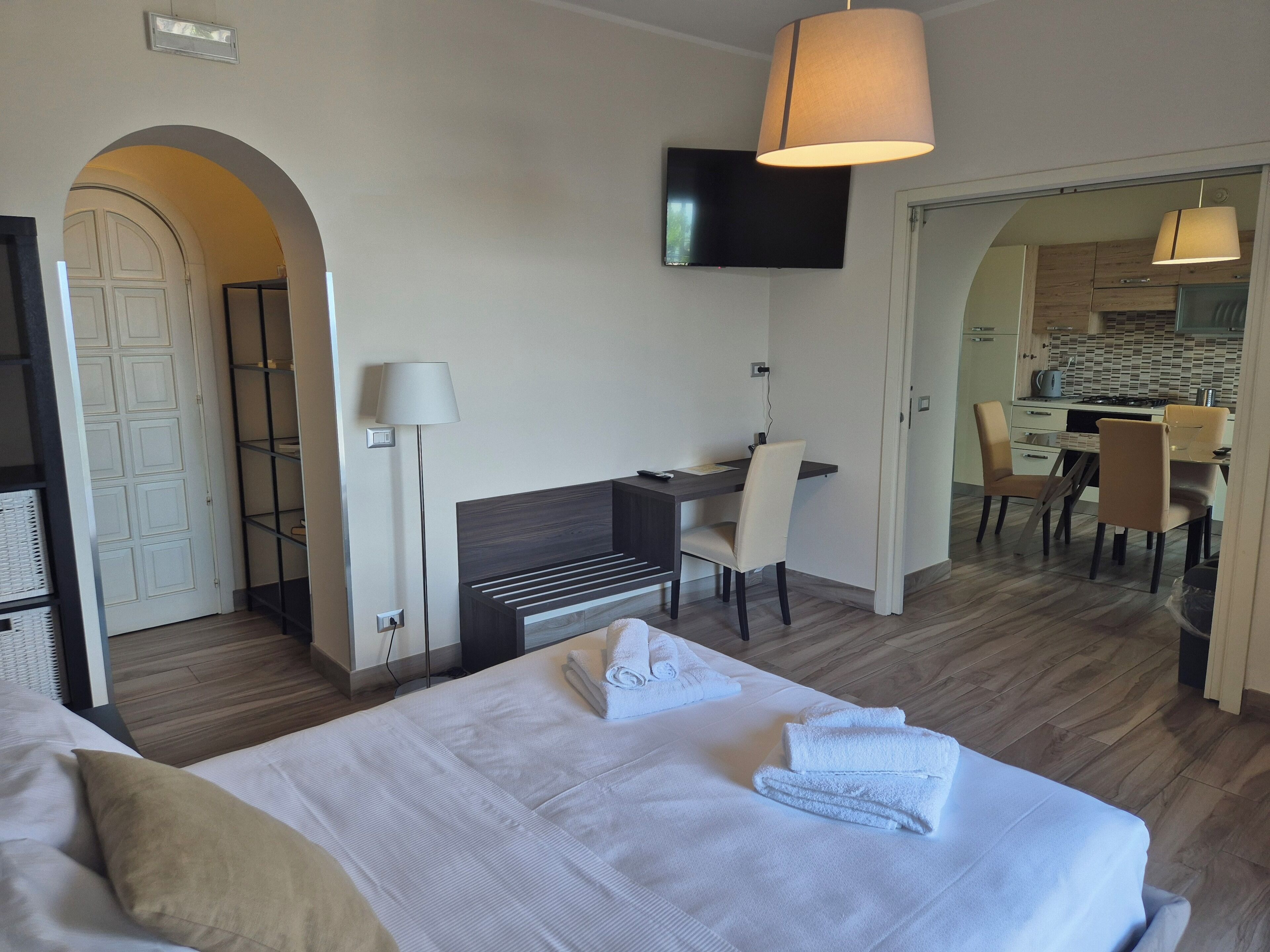 Habitación de hotel con cama, escritorio, silla, televisor y área de cocina visible a través de una puerta abierta.