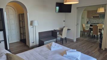 Habitación de hotel con cama, escritorio, silla, televisor y área de cocina visible a través de una puerta abierta.
