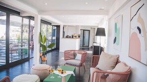 Habitación moderna y luminosa con muebles de mimbre, un reposapiés verde y una mesa con bebidas. Amplias ventanas ofrecen vistas al exterior.