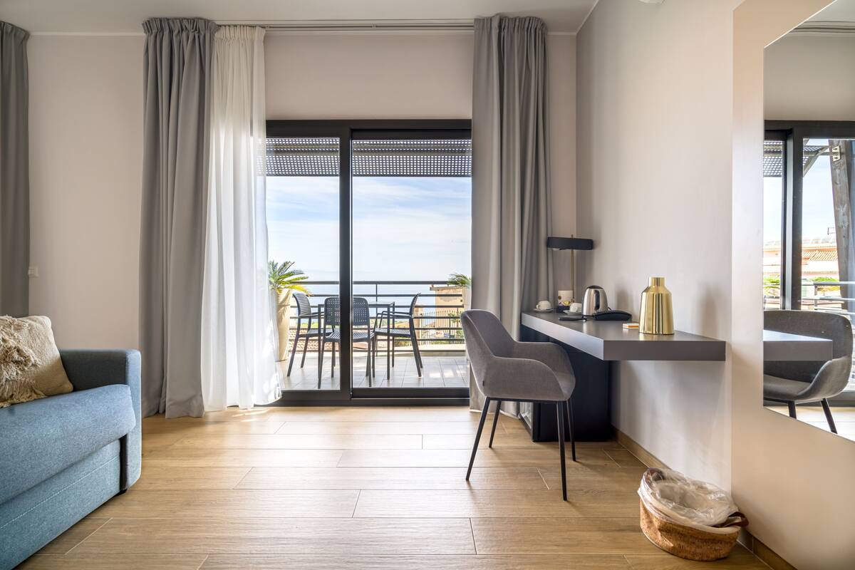 A modern hotel room with a balcony, a sofa, a desk with chairs, and a view of the cityscape.