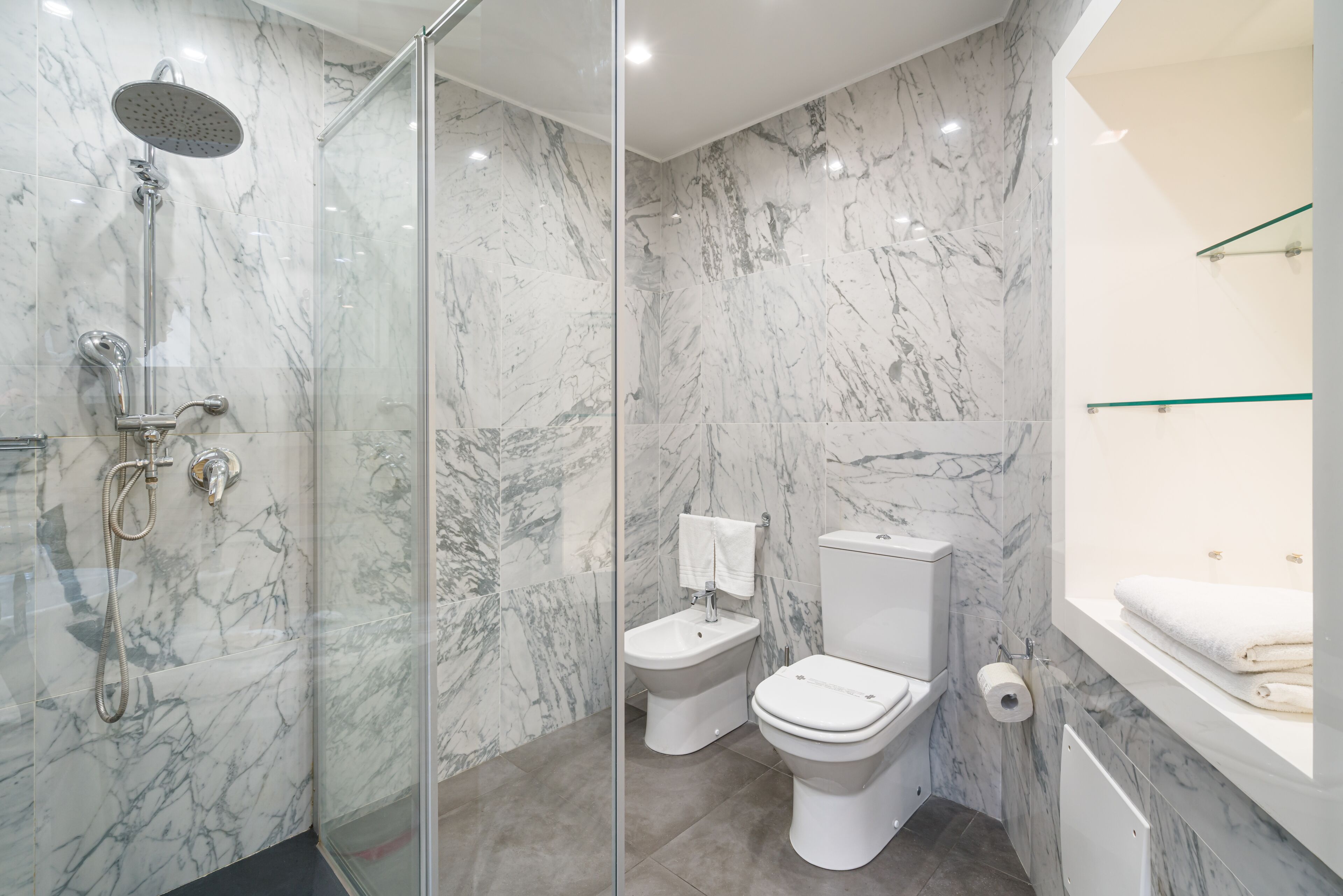 Une salle de bain moderne dotée d’une douche à parois de verre, d’un bidet, de toilettes et d’une baignoire avec une étagère en verre.