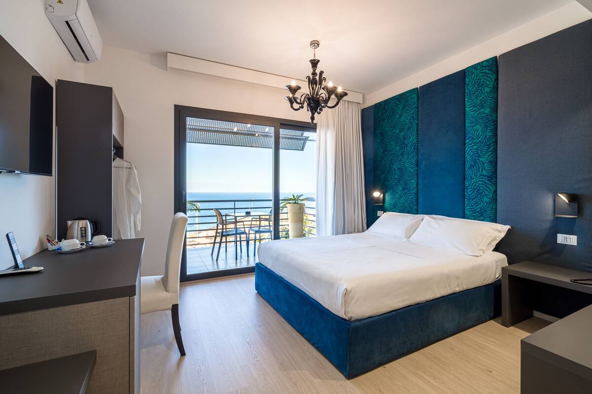 A modern hotel room with a large bed, a desk, and a view of the sea.