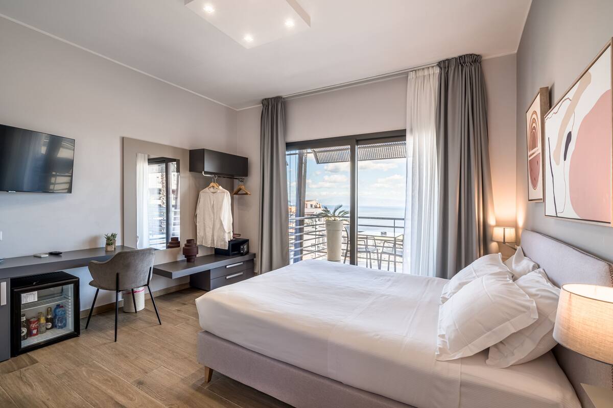 A modern hotel room with a large bed, a desk, a TV, and a balcony with a view.