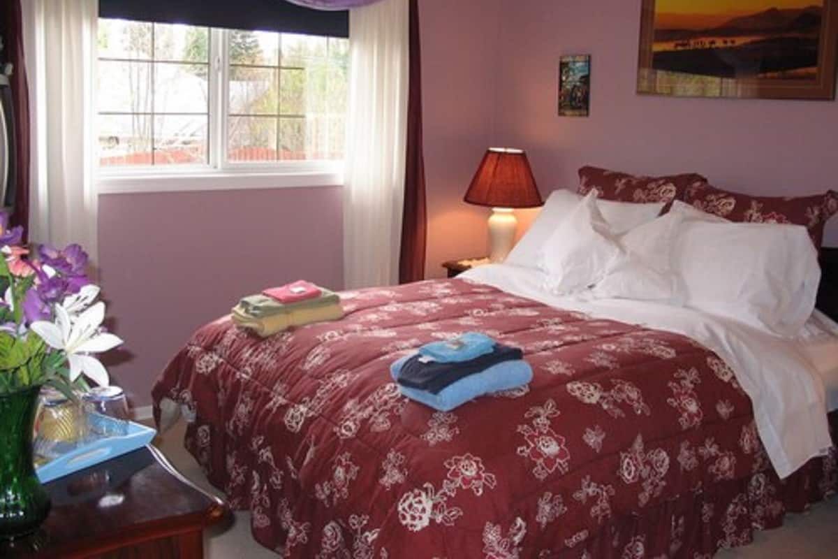 A bedroom with a bed, a nightstand, a lamp, a window with curtains, and a painting on the wall.