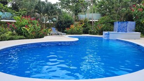Una piscina con borde de azulejos, rodeada de exuberante vegetación y un solárium con baldosas.