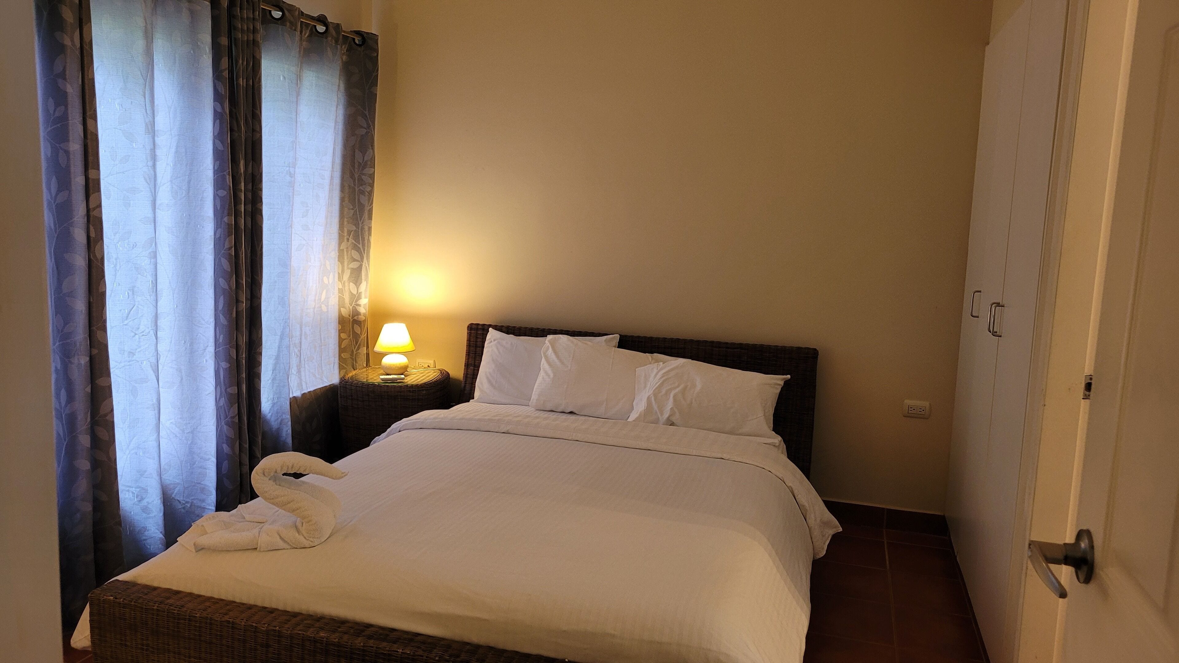 A neatly made bed with white linens and pillows, a bedside lamp, and a wicker chair.