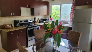 A kitchen with dark wooden cabinets, a stainless steel refrigerator, a microwave, and a stove. There is a wicker chair and a glass table with a vase of flowers. - Corinto Pearl Eco Resort (El Porvenir)