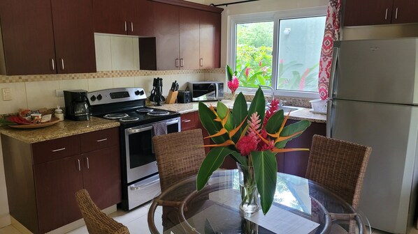A kitchen with dark wooden cabinets, a stainless steel refrigerator, a microwave, and a stove. There is a wicker chair and a glass table with a vase of flowers.