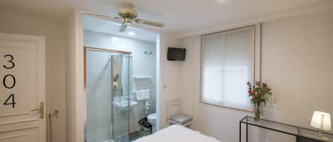 A hotel room with a bed, a glass-enclosed shower, a television, and a window with blinds.