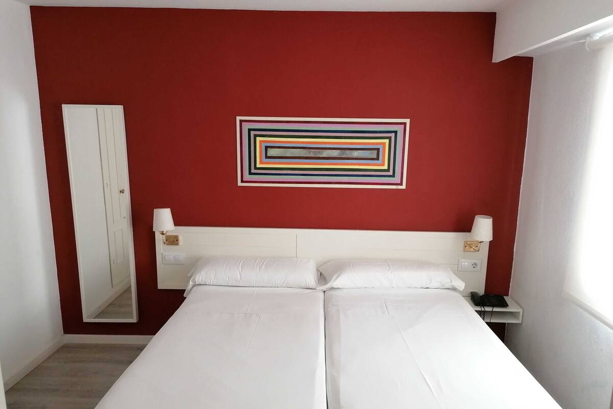 A hotel room with two single beds, a red accent wall, a mirror, a bedside table with a lamp, and a phone.
