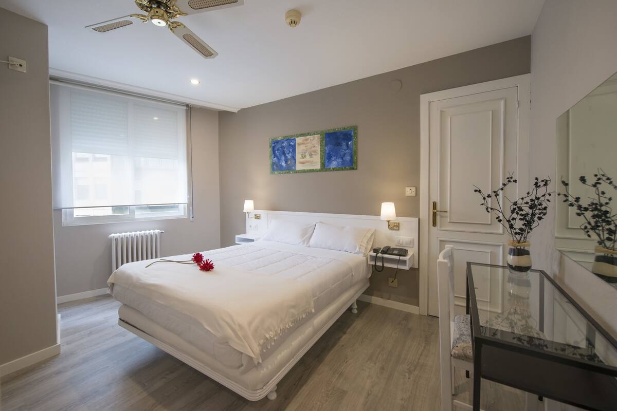 A modern bedroom with a large bed, bedside tables, a glass table, and a window with blinds.