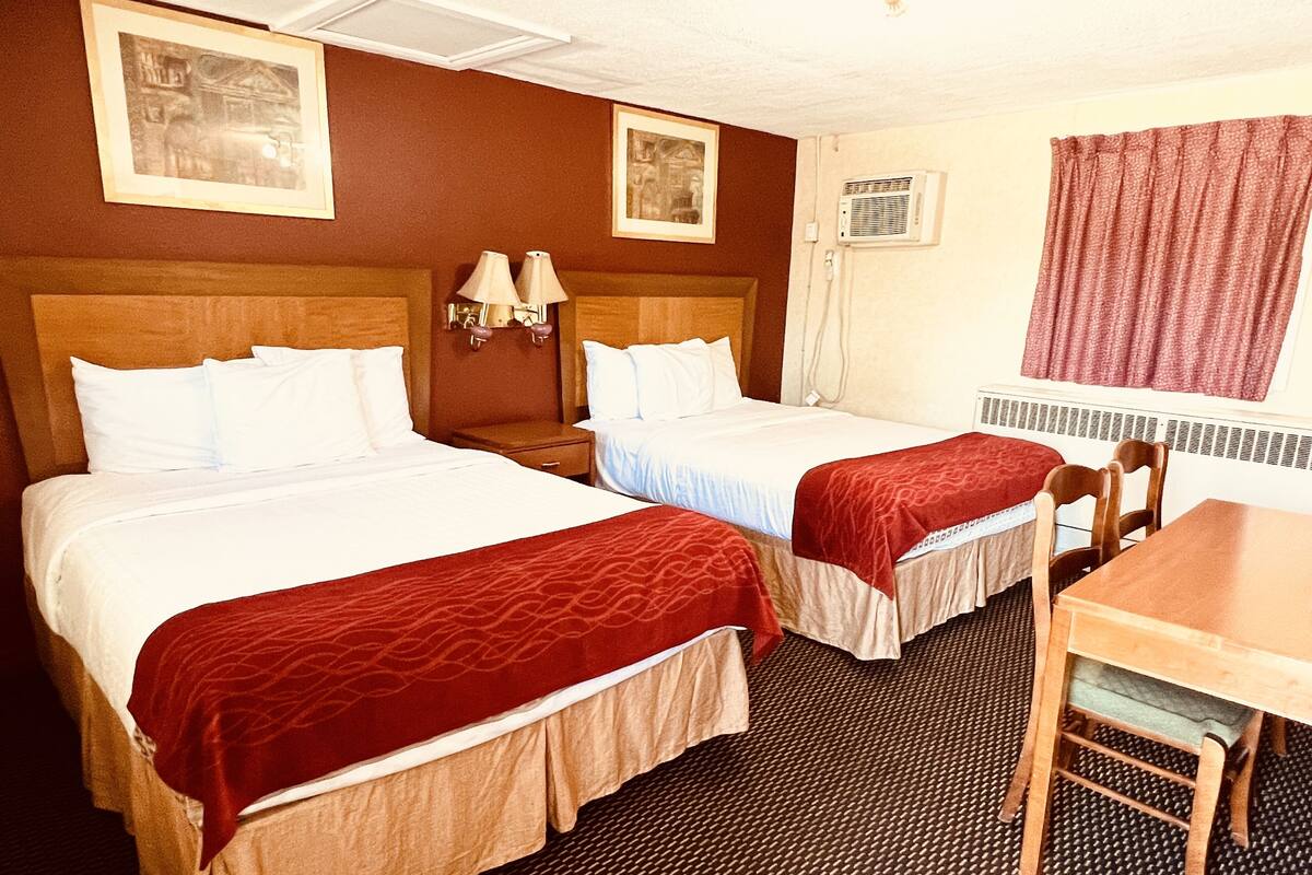 A hotel room with two beds, a small table, and a chair. There are two wall-mounted lamps and a window with curtains.