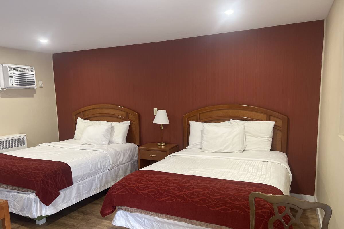 A hotel room with two beds, each with white bedding and red throw blankets. There is a wooden nightstand with a lamp between the beds, and a wooden chair in the corner.