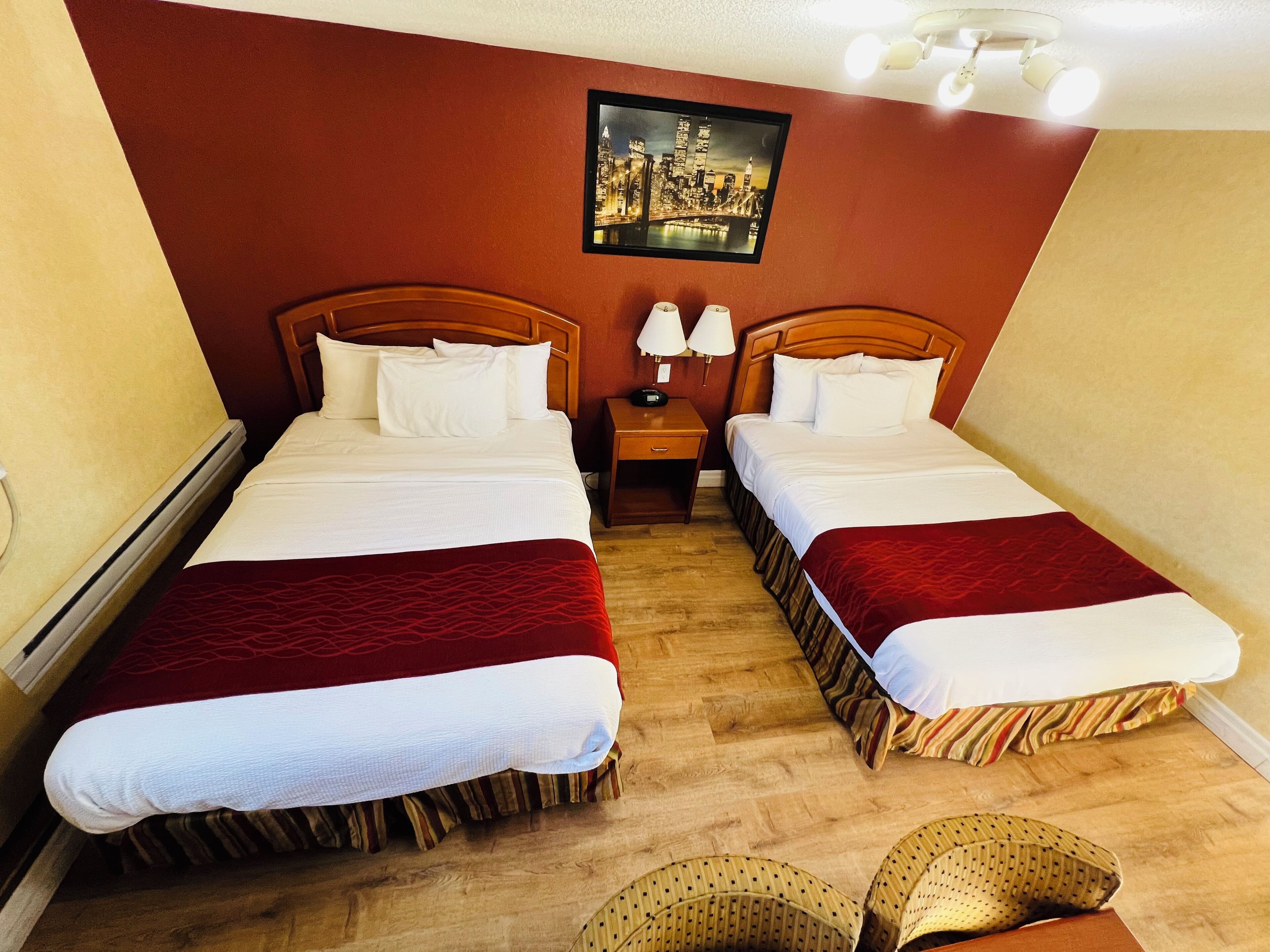 Habitación de hotel con dos camas, cabeceros de madera, ropa de cama blanca, pared de acento roja, un cuadro con una vista urbana y una mesita de noche con dos lámparas.