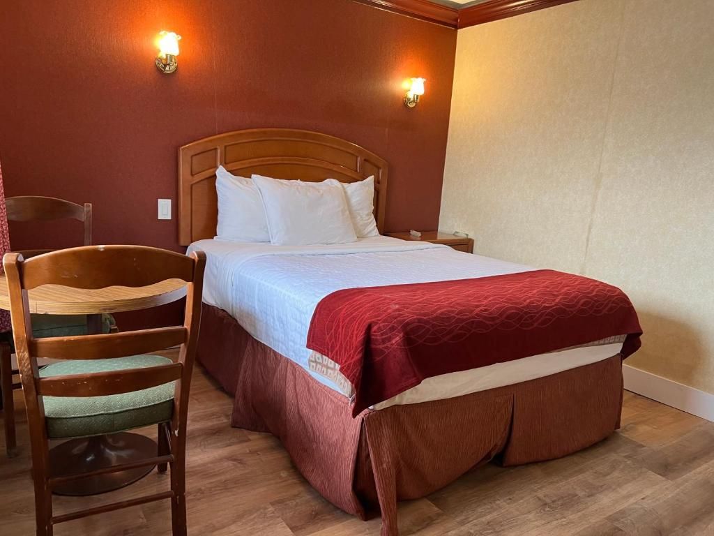Quarto de hotel com cabeceira de madeira, cama com roupa de cama branca e detalhes vermelhos, uma cadeira de madeira e duas luminárias fixadas na parede.
