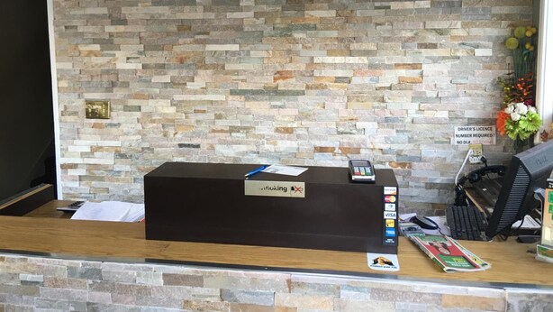 A reception desk with a stone wall backdrop, a nameplate, a computer, a telephone, and a magazine. - Bestway Motel (Windsor)