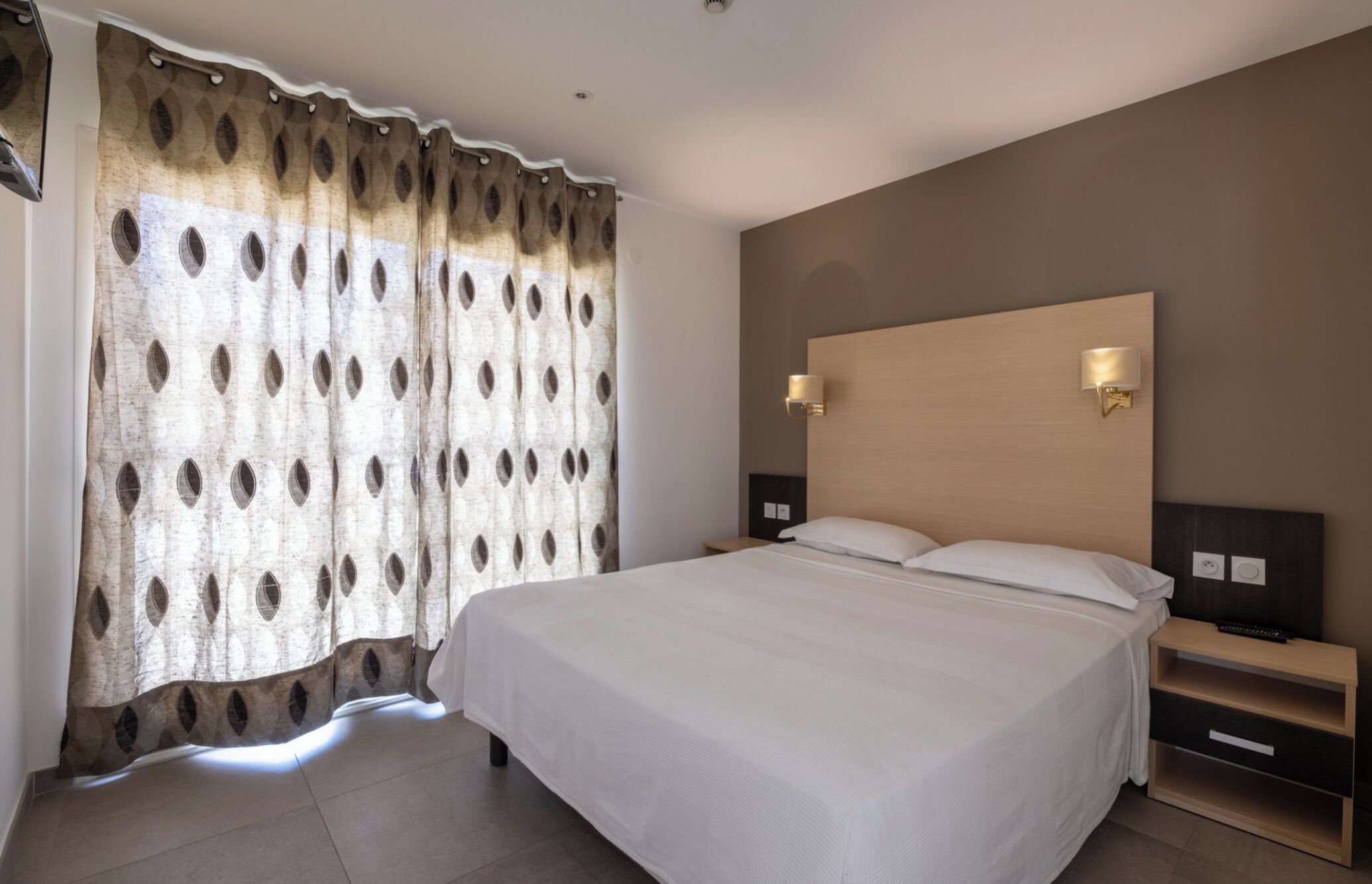 Quarto de hotel com uma cama grande, duas mesas de cabeceira e cabeceira com luzes fixadas na parede.