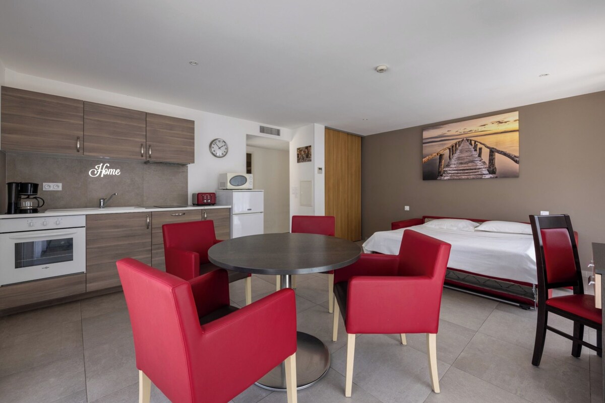 A compact hotel room with a kitchenette, a dining area with red chairs, a bed, and a painting of a pier.