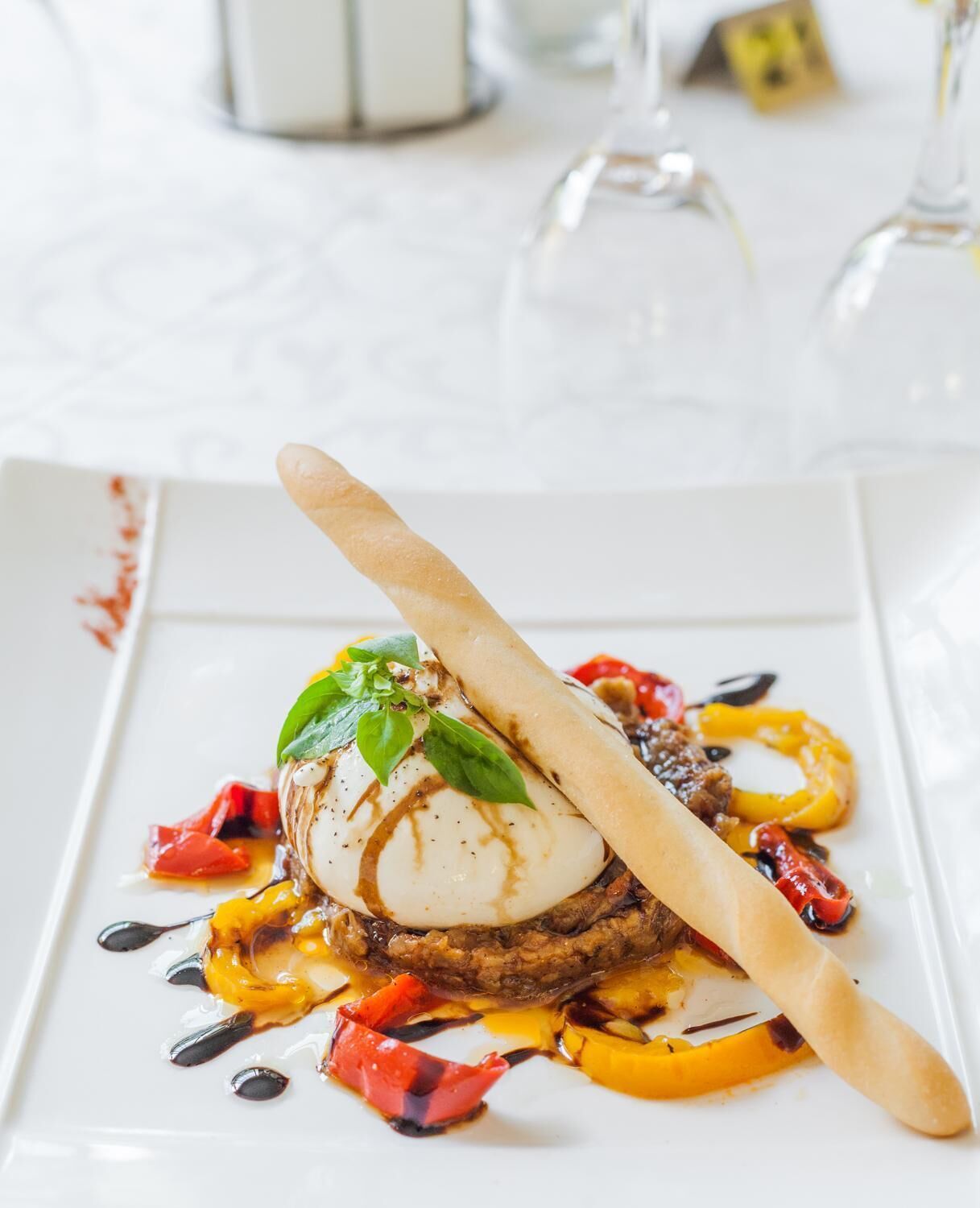 A white plate with a dish consisting of a poached egg, a piece of meat, and vegetables, garnished with basil and sauce.