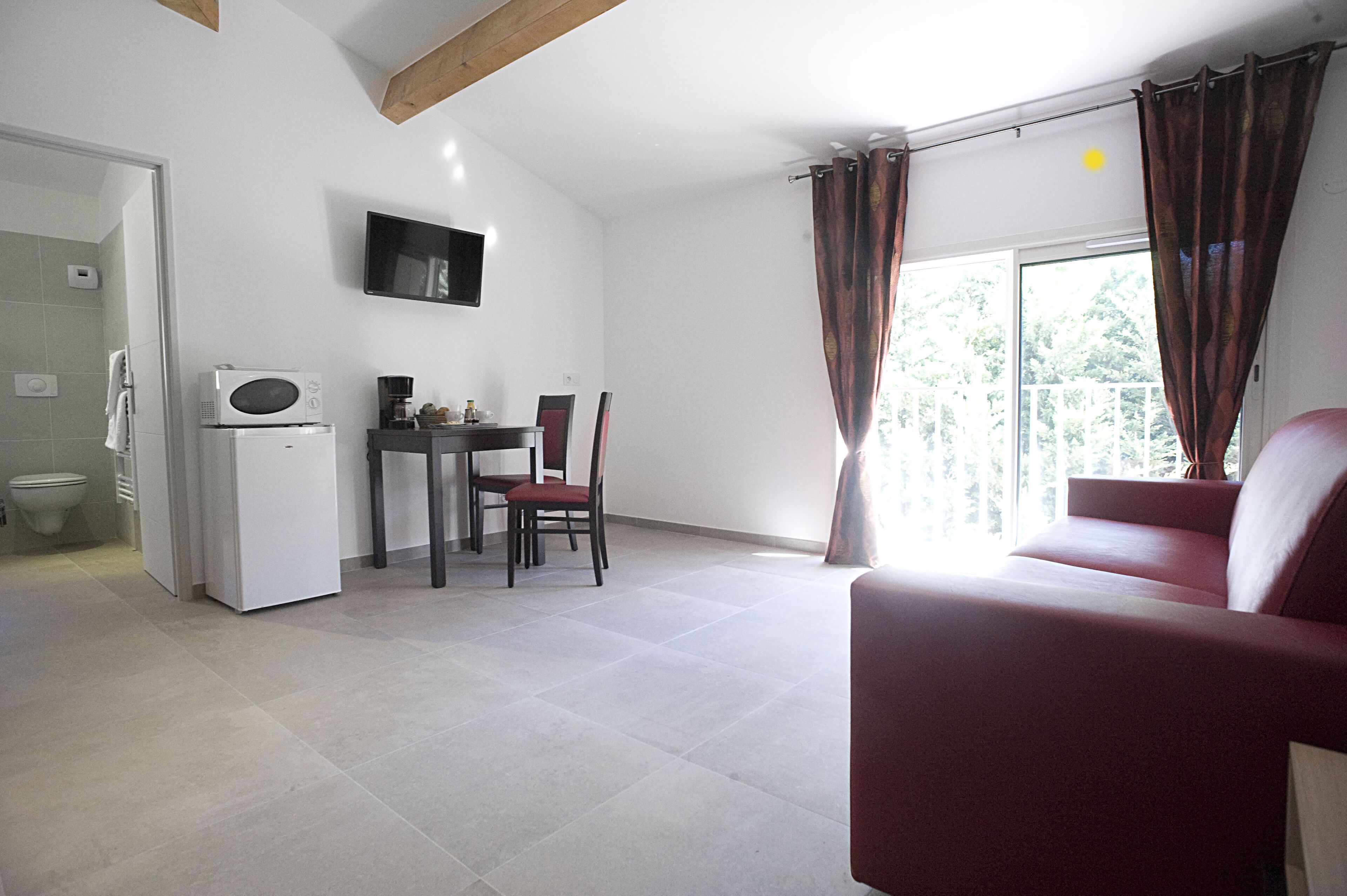 Ein Wohnzimmer mit einem roten Sofa, ein Essbereich mit Tisch und Stühlen, eine Küche mit Mikrowelle und Kühlschrank sowie ein Balkon mit Vorhängen.