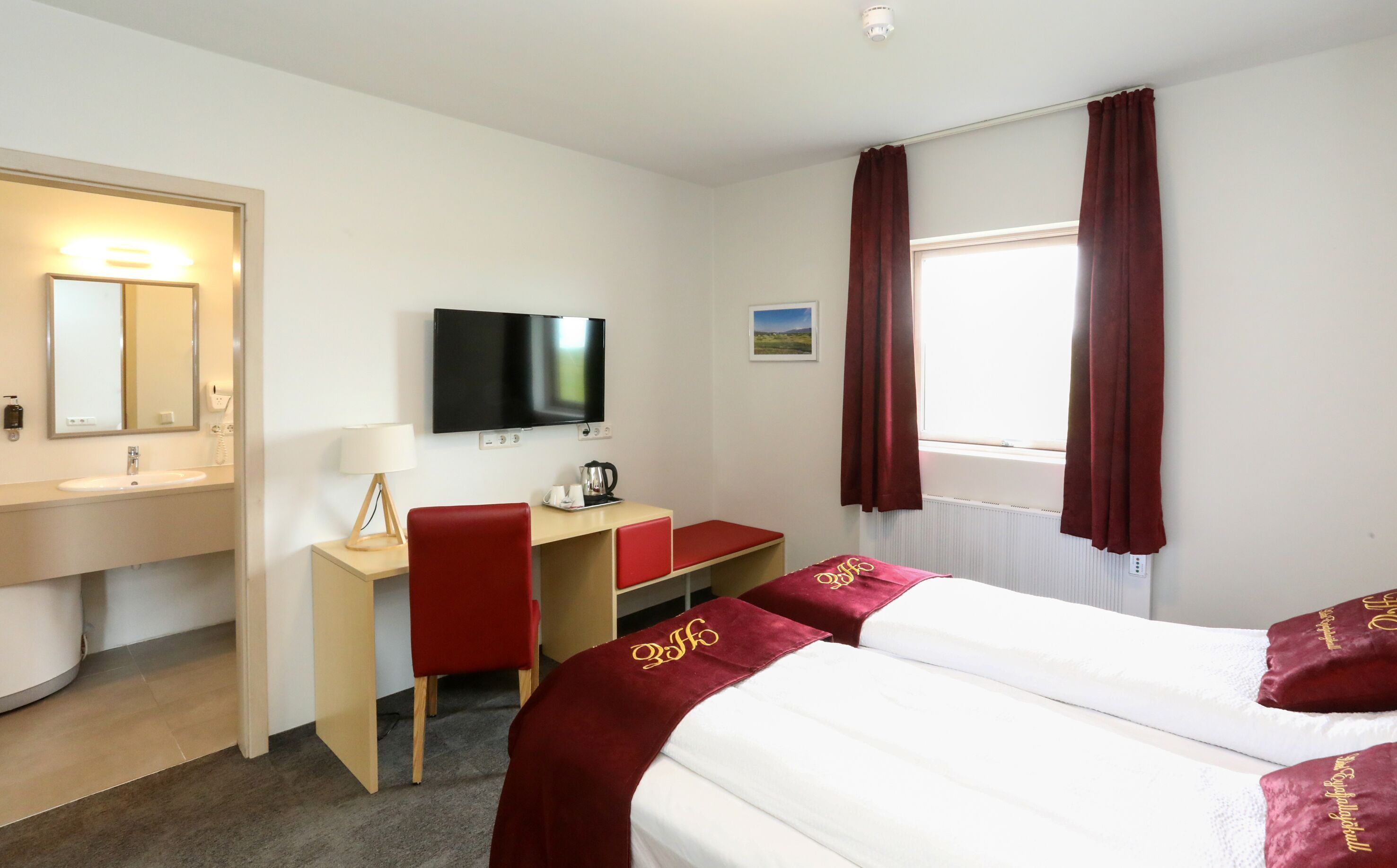 Une chambre d’hôtel comprenant un lit, un bureau avec une chaise, une télévision, une fenêtre avec des rideaux, un lavabo et un miroir.