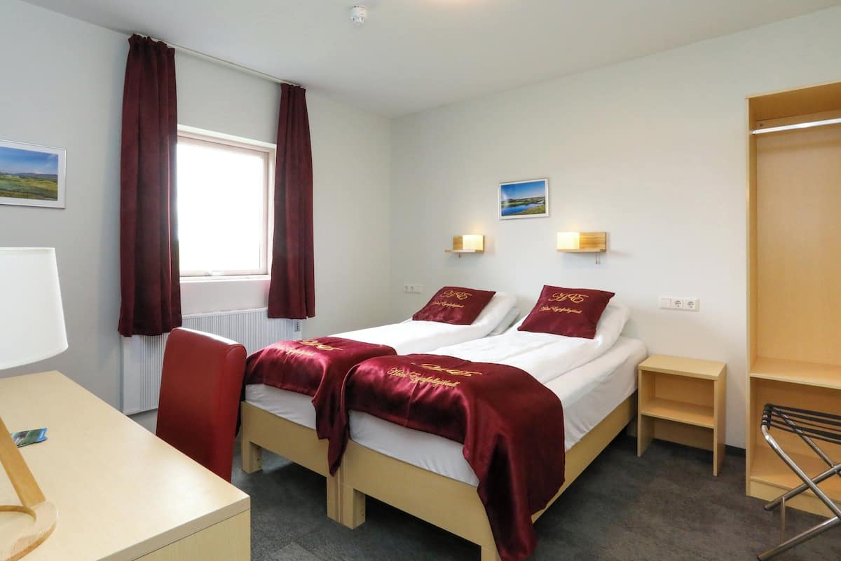 Ein Hotelzimmer mit zwei Betten, einem Schreibtisch, einem Stuhl, einem Fenster mit Vorhängen, Wandleuchten und Blick auf eine Landschaft.