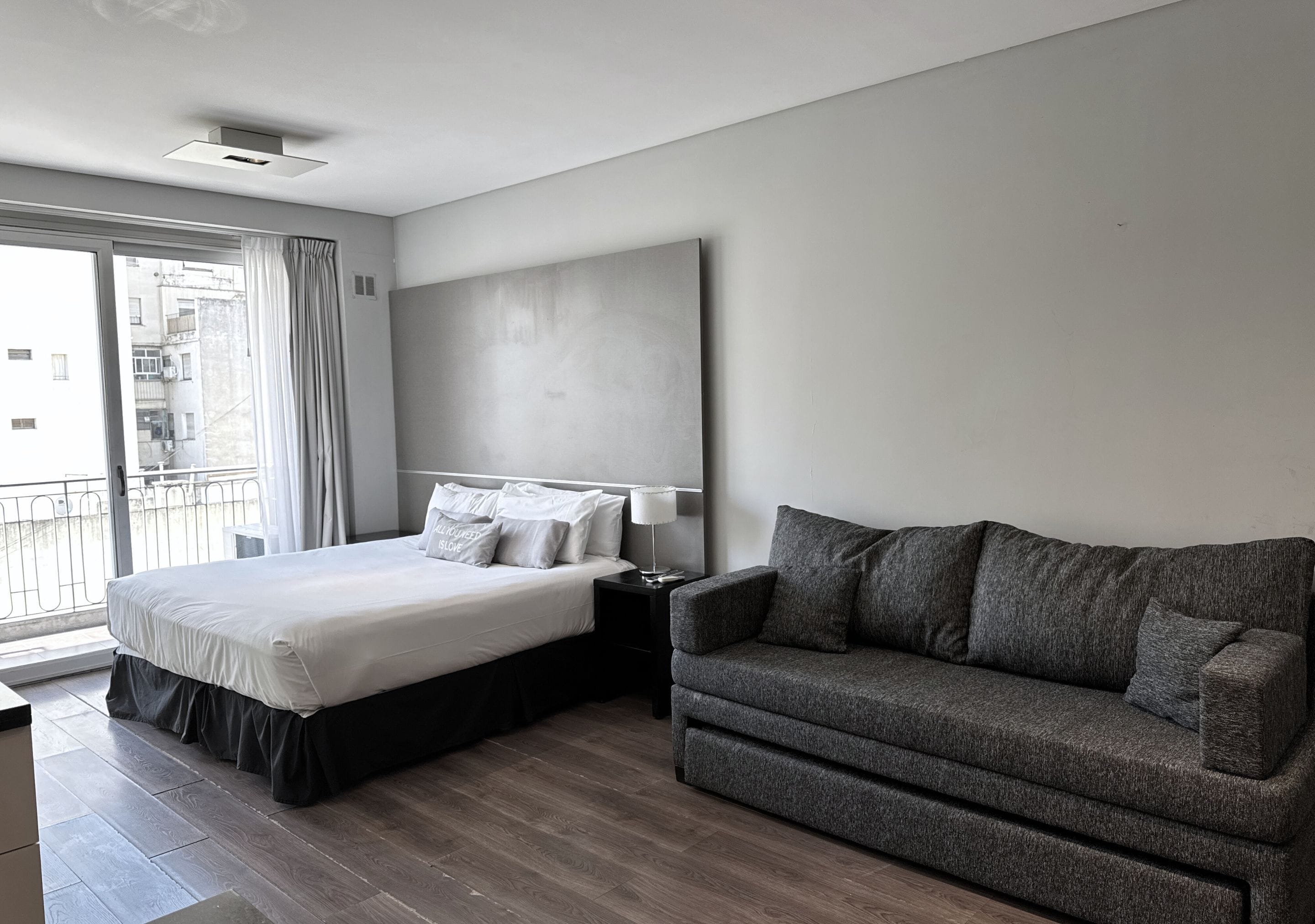 A modern hotel room with a large bed, a grey sofa, and a large window offering a view of the city.
